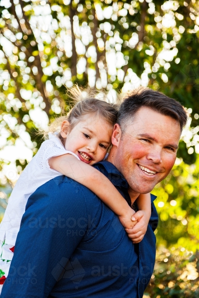 Happy parent giving child piggyback outside smiling as they play together - Australian Stock Image