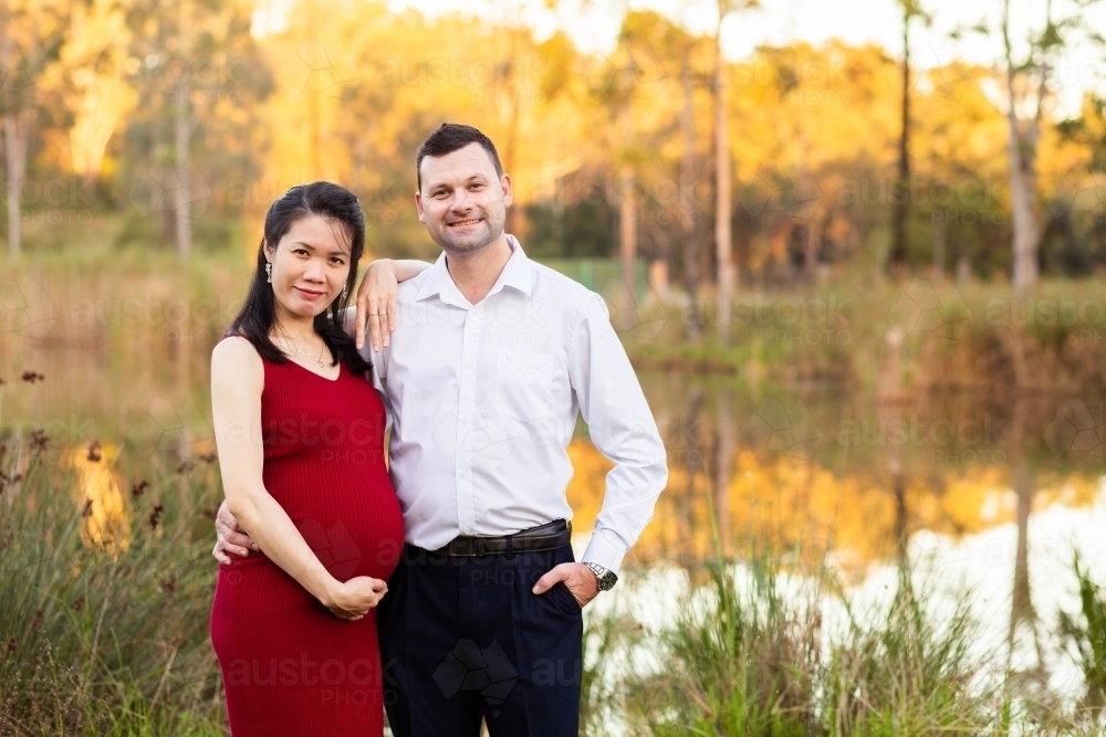 Happy pregnant interracial Aussie couple standing outside near dam - Australian Stock Image