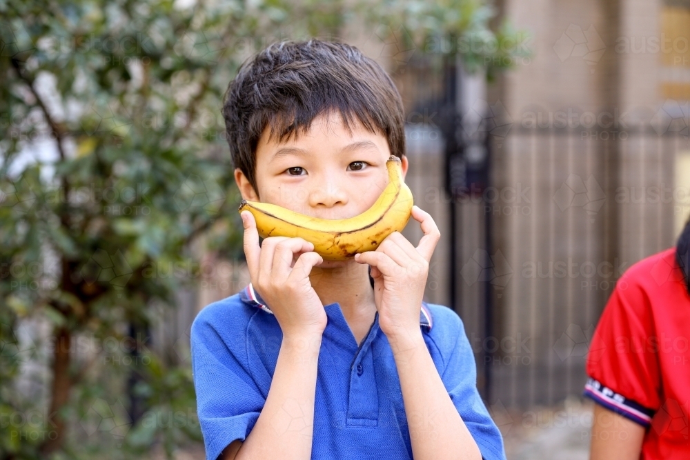 Happy primary school student holding a banana up to his face - Australian Stock Image