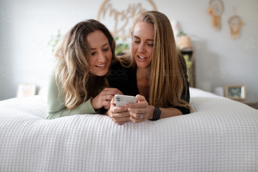 Happy same sex couple lying on a bed looking at a smart phone - Australian Stock Image