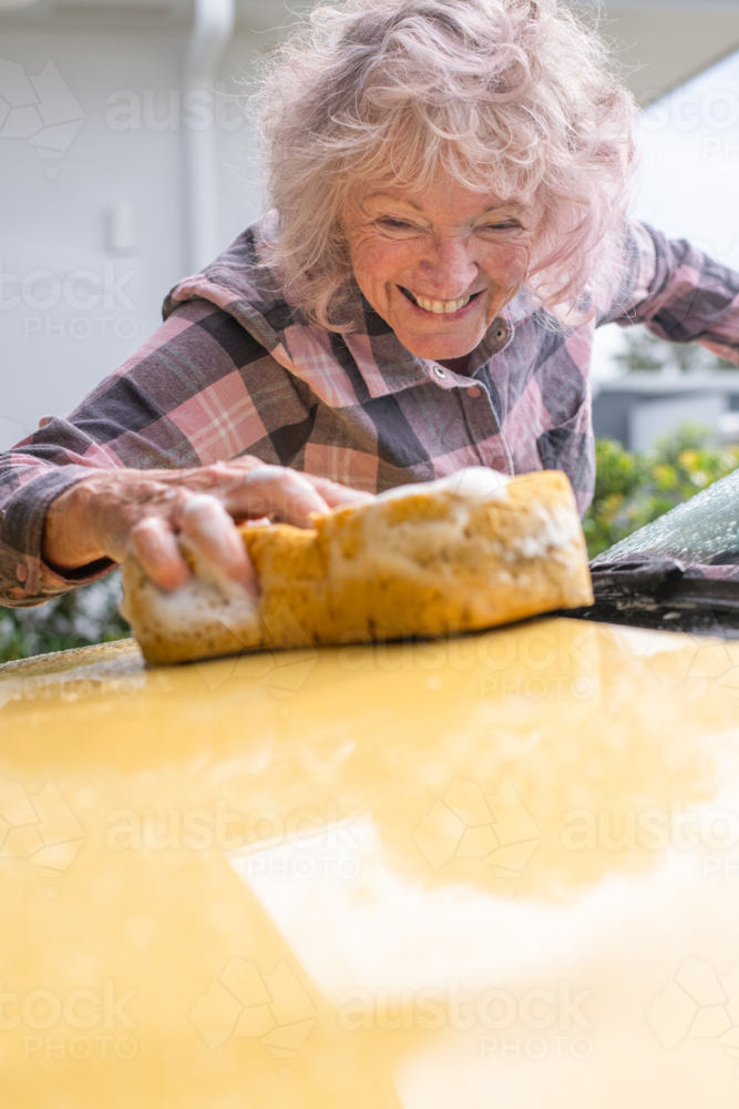 happy senior woman washing he car with a large yellow sponge - Australian Stock Image