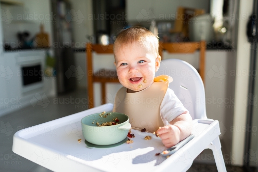 Happy seven month old baby eating breakfast in highchair in morning sunlight - Australian Stock Image