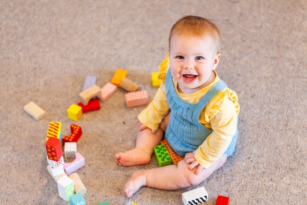 Happy seven month old baby playing with coloured blocks on carpet floor - Australian Stock Image