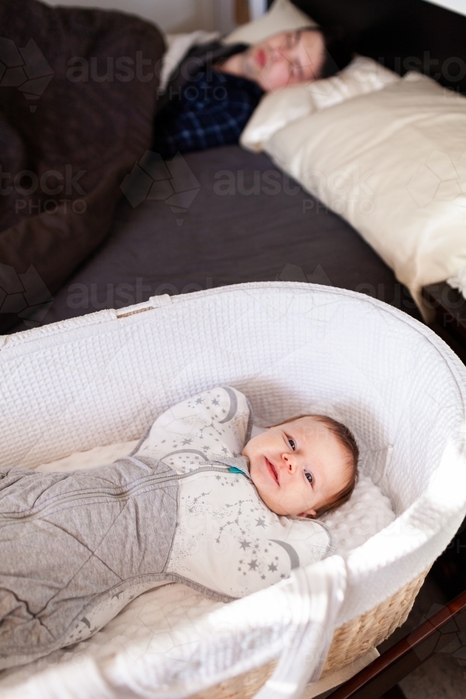 happy smiling baby wrapped in swaddle awake in bassinet in morning with parent sleeping in bed - Australian Stock Image