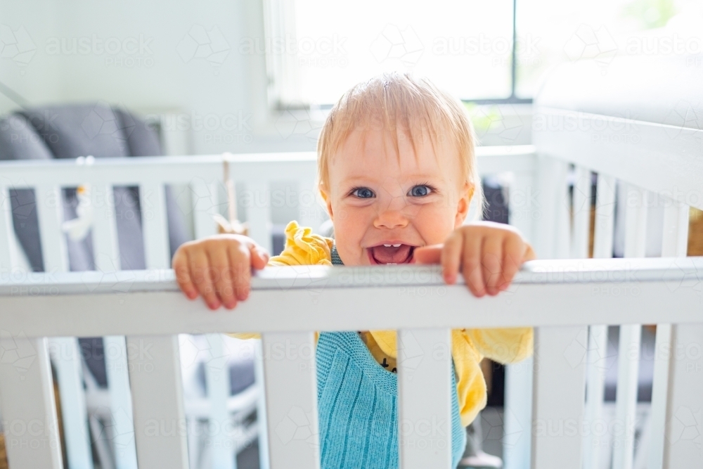 happy smiling child standing up in crib in bedroom - Australian Stock Image
