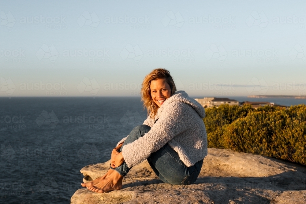 Happy smiling young woman sitting on coastal clifftops at sunrise - Australian Stock Image
