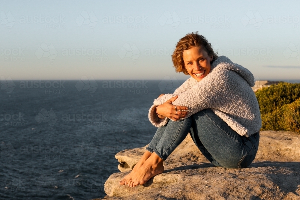 Happy smiling young woman sitting on coastal clifftops at sunrise - Australian Stock Image