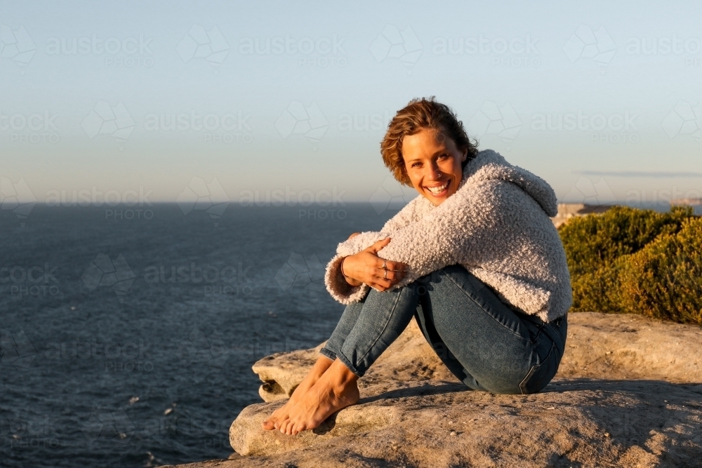 Happy smiling young woman sitting on coastal clifftops at sunrise - Australian Stock Image