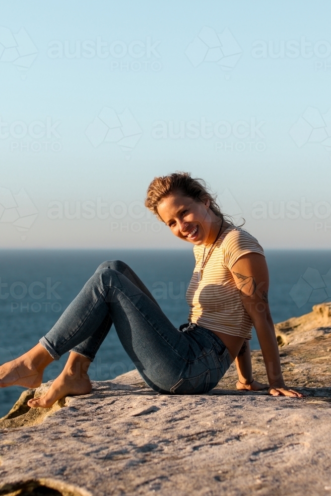 Happy smiling young woman sitting on coastal clifftops at sunrise - Australian Stock Image
