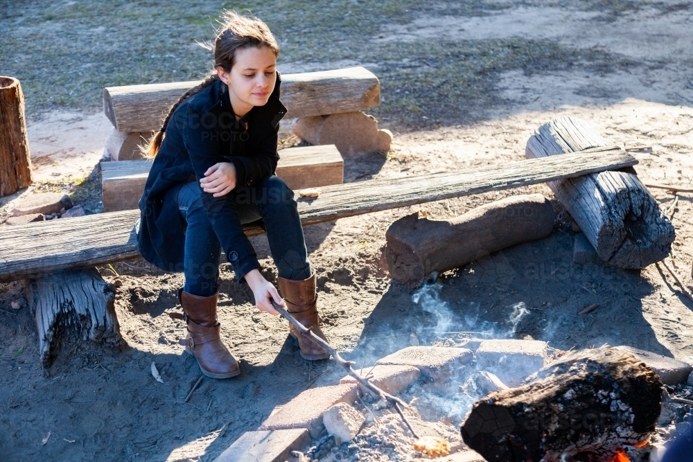 Happy teen girl poking campfire with a stick on winter morning - Australian Stock Image