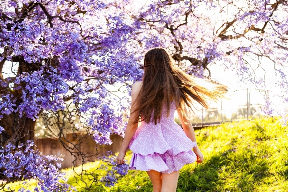 Happy teen girl with long hair and purple dress spinning and twirling beside flowering jacaranda - Australian Stock Image