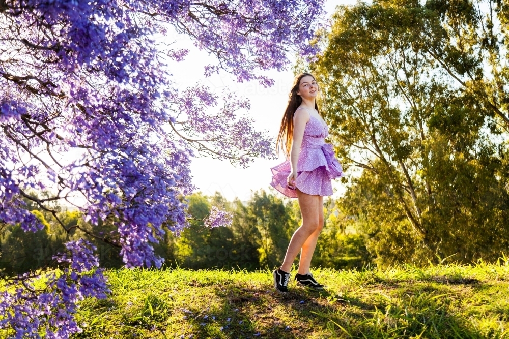 Happy teen girl with long hair and purple dress spinning and twirling beside flowering jacaranda - Australian Stock Image