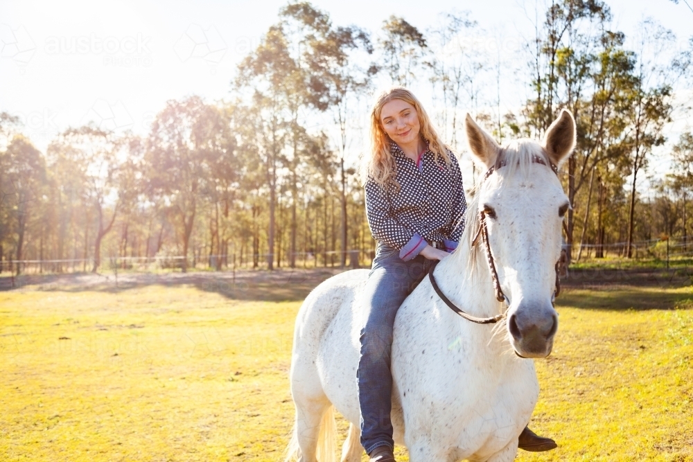 Happy teen person riding her horse in sunlit paddock - Australian Stock Image
