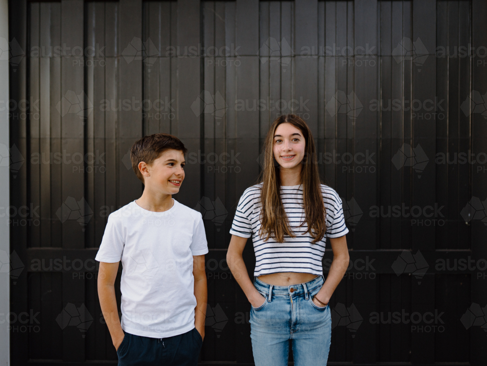 happy teen siblings standing outside against black garage door background - Australian Stock Image