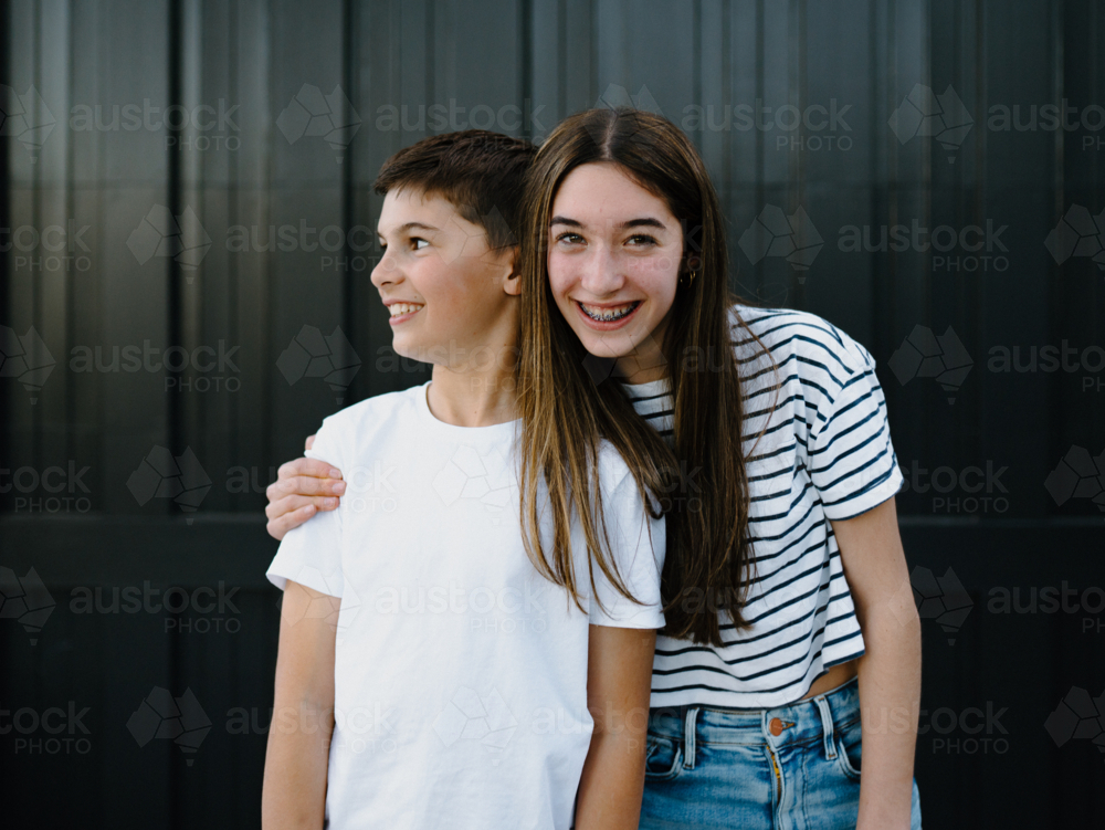 happy teen siblings standing outside against black garage door background - Australian Stock Image