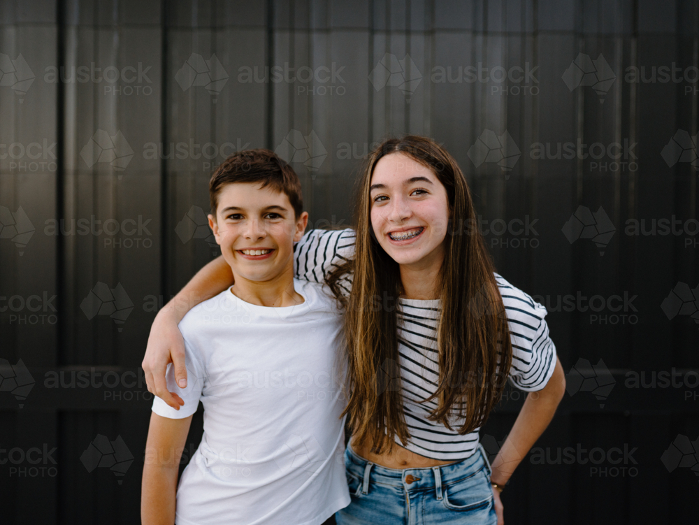 happy teen siblings standing with arms on each others shoulders against black garage door - Australian Stock Image