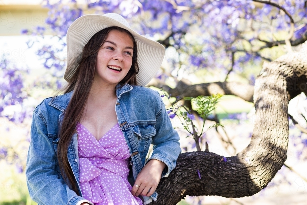 Happy teenage girl wearing a hat for sun protection sitting in a jacaranda tree laughing - Australian Stock Image
