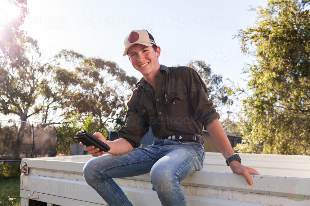 happy teenaged young man sitting on ute tray using mobile phone - Australian Stock Image