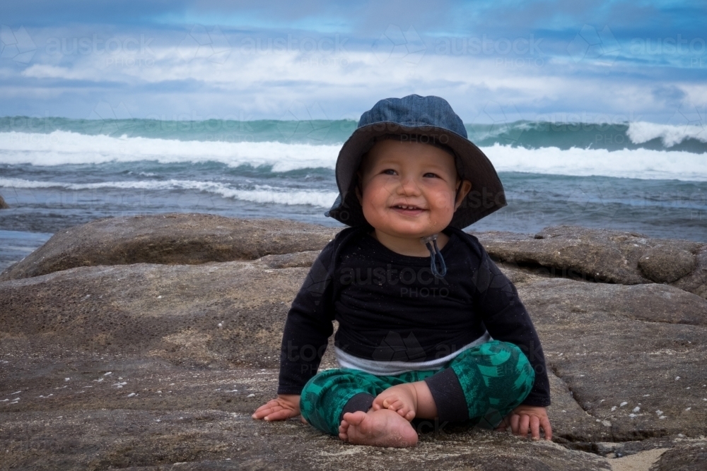 Happy toddler sitting on rocky beach with waves in background - Australian Stock Image