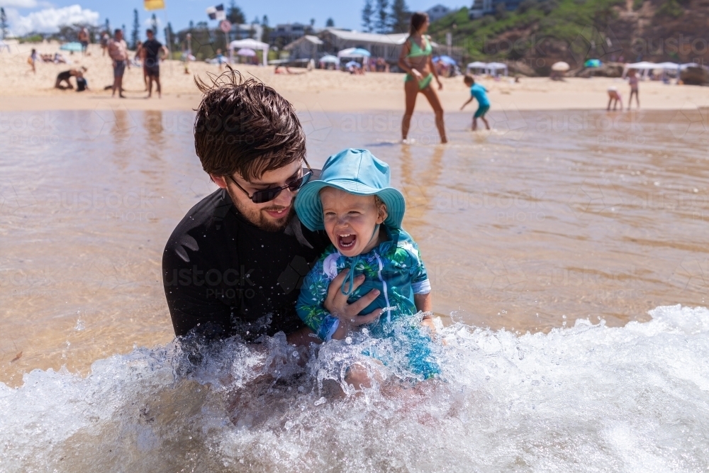 Happy toddler wearing hat at beach laughing in waves with parent - Australian Stock Image