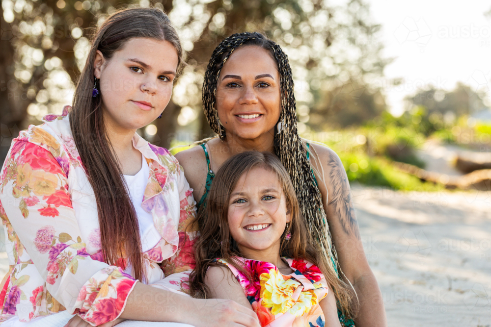 Happy Torres Strait Islander woman with her two girls sitting together outside on the coast - Australian Stock Image