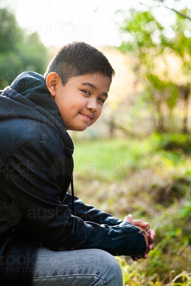 Happy tween boy sitting on log outside smiling - Australian Stock Image