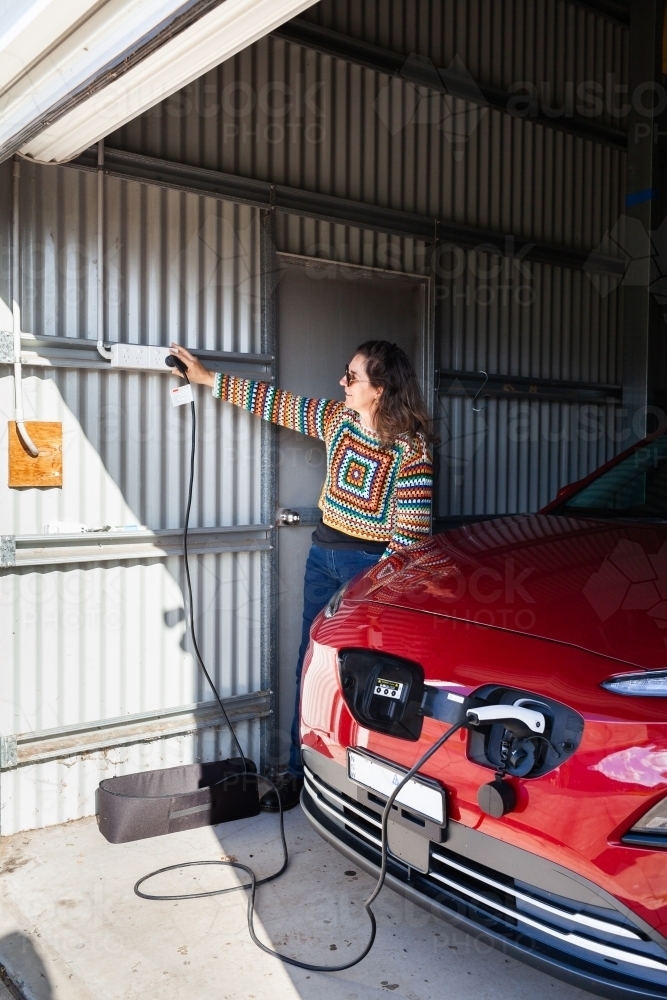 Happy woman in shed garage switching on the electric vehicle car charger at the wall - Australian Stock Image