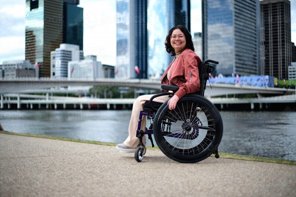 Happy woman in wheelchair in front of river with city in background - Australian Stock Image
