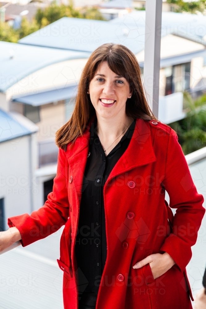 Happy woman standing on balcony in red jacket - Australian Stock Image