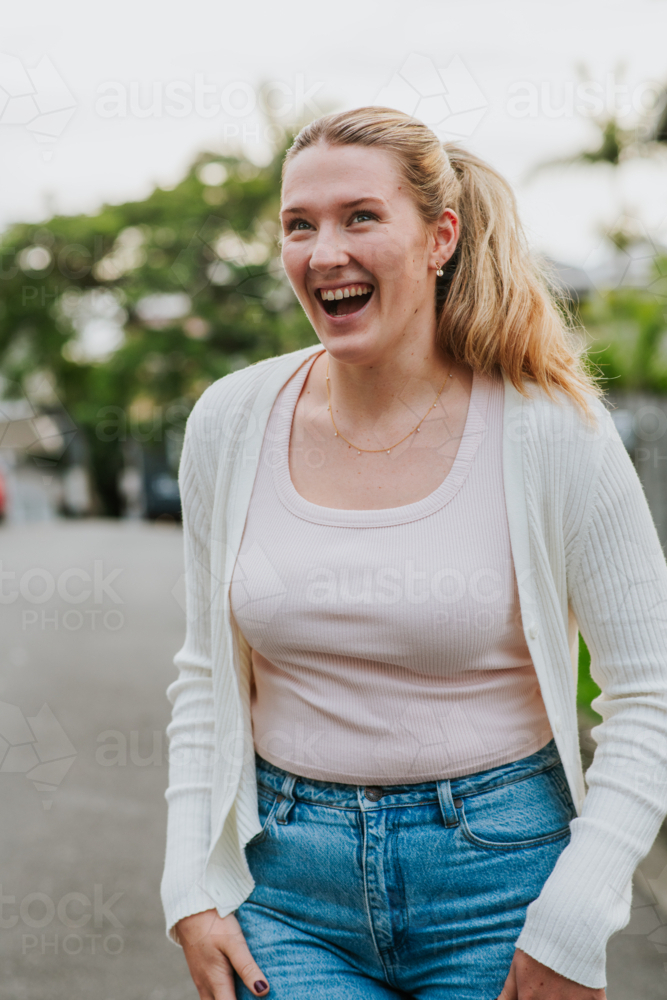 happy woman with a ponytail stands outdoors wearing a white cardigan, pink top, and blue jeans - Australian Stock Image