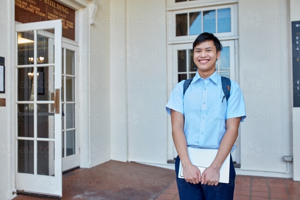 Happy young Asian male student standing holding laptop - Australian Stock Image