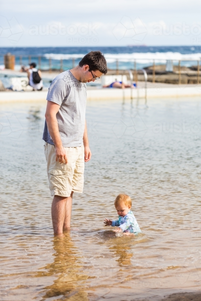 Happy young baby girl sitting in shallow sea water in ocean pool at beach in summer - Australian Stock Image
