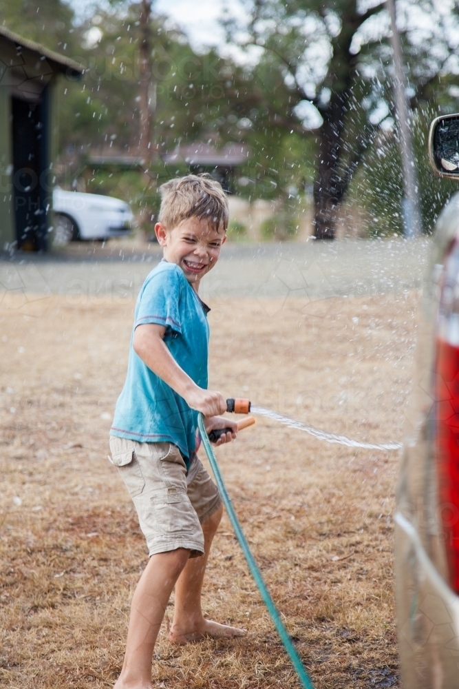 Happy young boy laughing and washing down family car with hose - Australian Stock Image