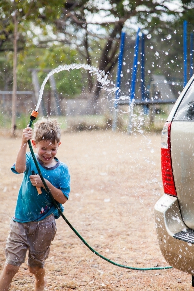 Happy young boy laughing and washing down family car with hose - Australian Stock Image