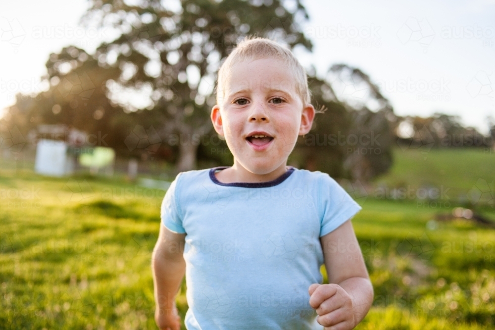 Happy young boy runs forward on green grass in golden afternoon light. - Australian Stock Image