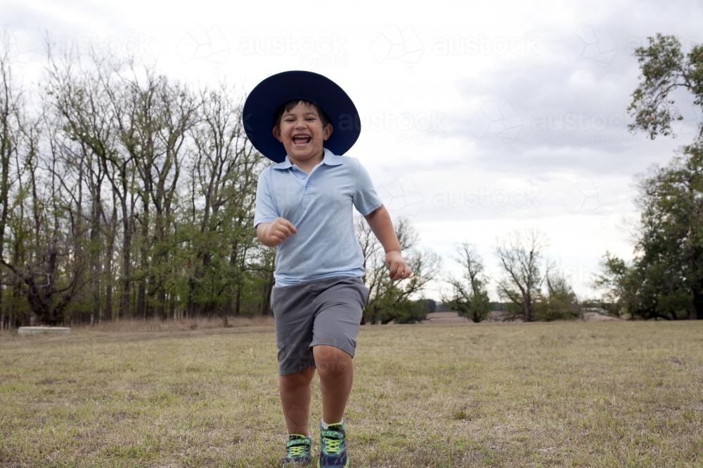 Happy young boy wearing school uniform running across grass - Australian Stock Image
