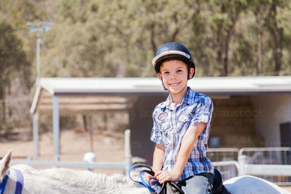 Happy young boy with safety horse riding helmet on horseback during lesson - Australian Stock Image