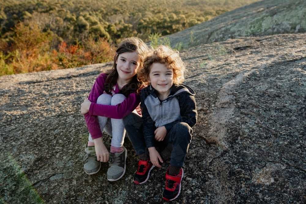 Happy young brother and sister sitting together on a rocky ledge in the Australian bush at sunset - Australian Stock Image