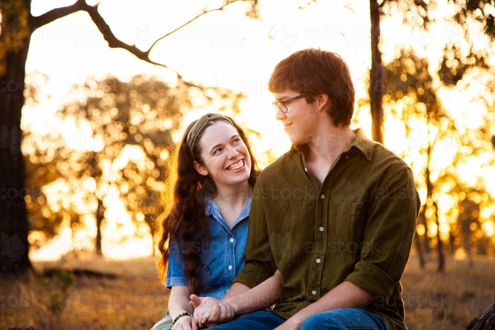 Happy young couple sitting on log at sunset smiling together - Australian Stock Image