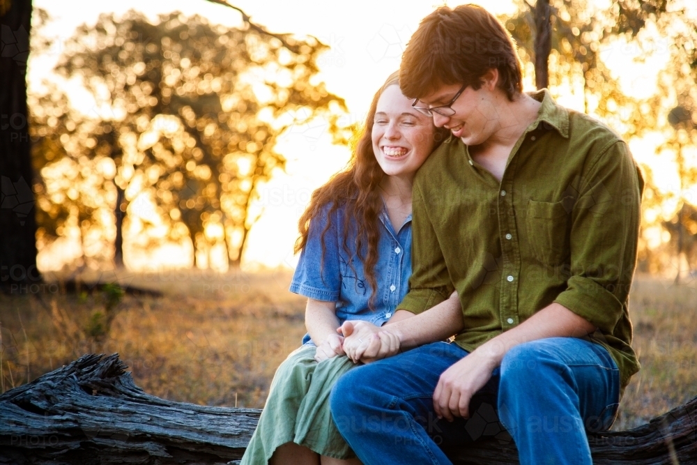 Happy young couple sitting on log at sunset smiling together - Australian Stock Image