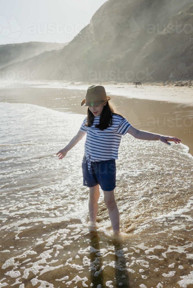 Happy young girl wearing a sunhat standing in the water at the beach in the afternoon - Australian Stock Image