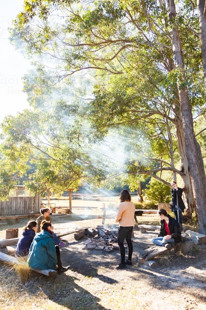 Happy young group of friends sitting around a campfire on a campground in the morning - Australian Stock Image