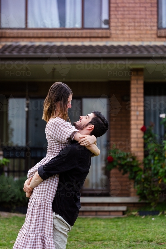 Happy young homebuyers - husband picking up wife in front of their house - Australian Stock Image