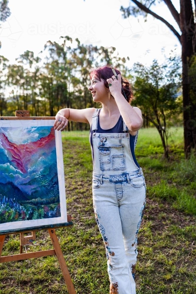 Happy young lady artist with paintbrush tucking hair behind ear - Australian Stock Image