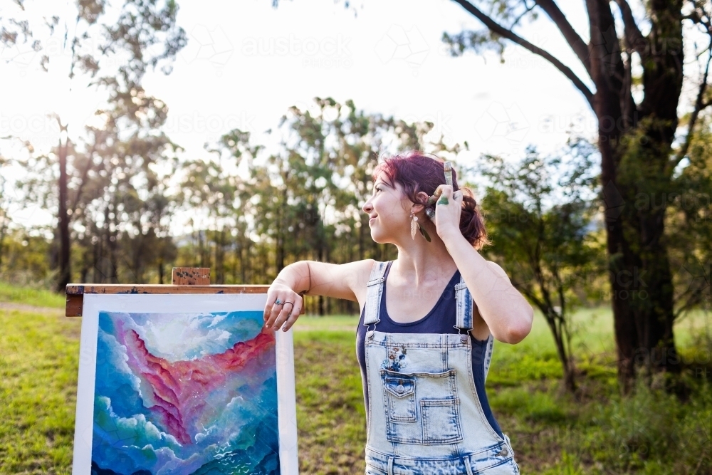 Happy young lady artist with paintbrush tucking hair behind ear - Australian Stock Image