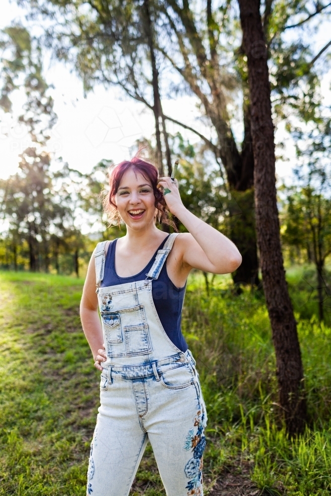 Happy young lady artist with paintbrush tucking hair behind ear - Australian Stock Image