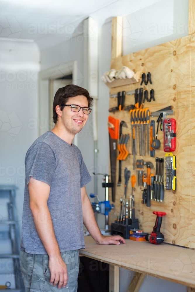 happy young man in home shed leaning on workbench with tools on board behind him - Australian Stock Image
