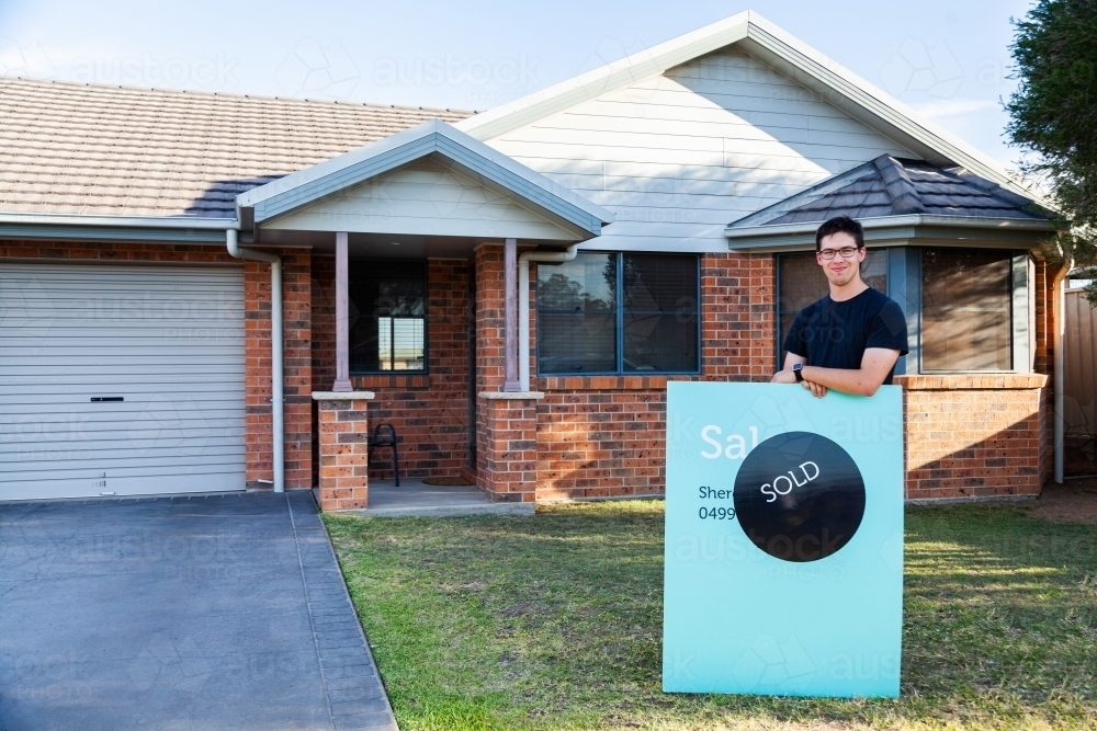 Happy young man outside of home with for sale sign - Australian Stock Image