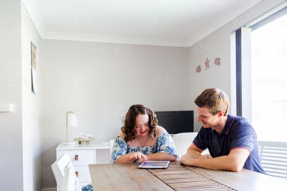 Happy young person using tablet device with disability worker at dining table - Australian Stock Image