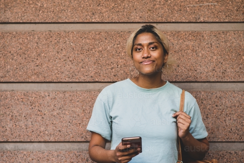 happy young woman leading against wall with mobile phone - Australian Stock Image